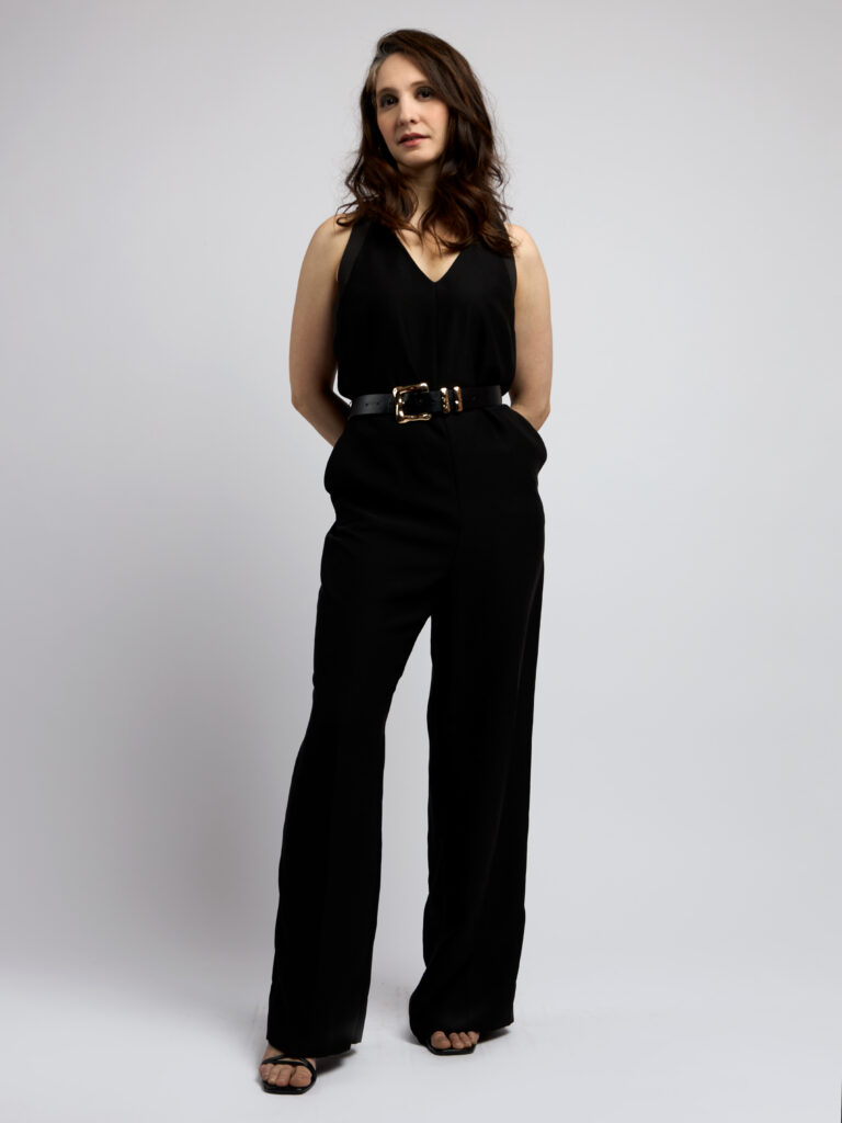 Full-length studio shot of a female model in a black sleeveless jumpsuit with a gold-buckle belt against a light backdrop