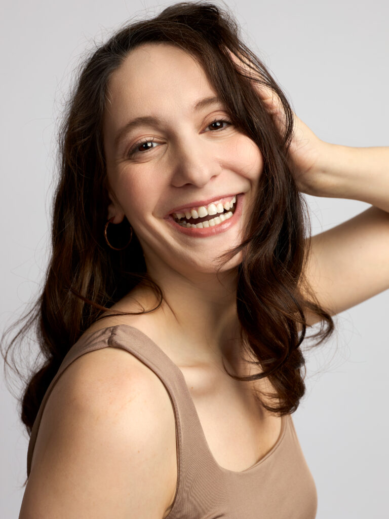 Brunette female model smiling and laughing in a beige tank top against a light grey studio backdrop