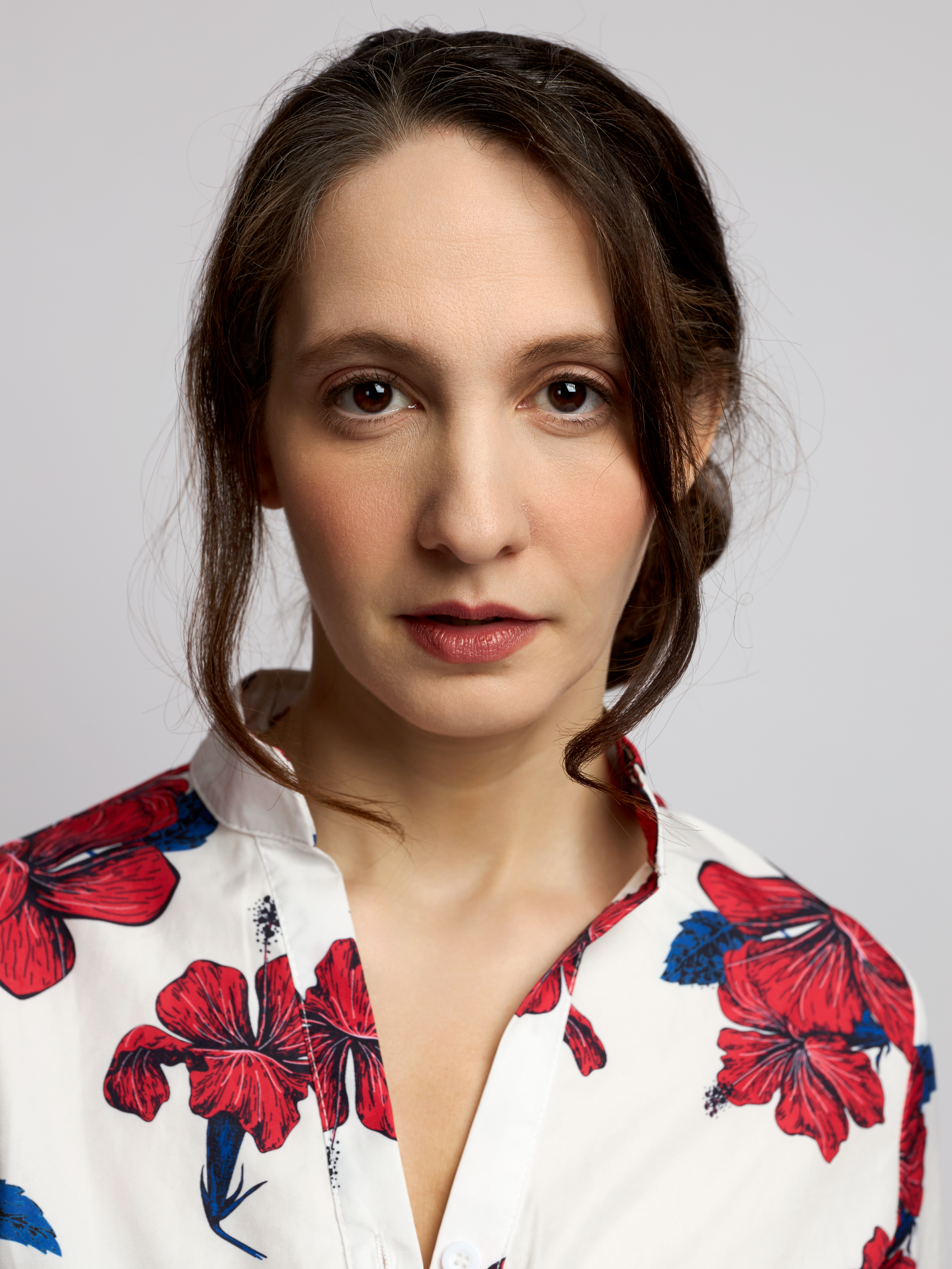 Close-up portrait of a brunette female model with a calm, direct expression wearing a white blouse with red and blue floral print